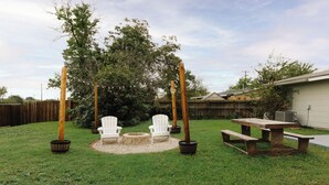 Un jardin avec une table de pique-nique en bois, deux fauteuils Adirondack, un foyer en pierre et deux poteaux en bois décorés de guirlandes lumineuses.