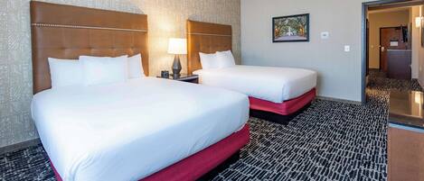 A hotel room with two beds, a patterned carpet, a headboard, a nightstand, a lamp, and a framed picture on the wall.
