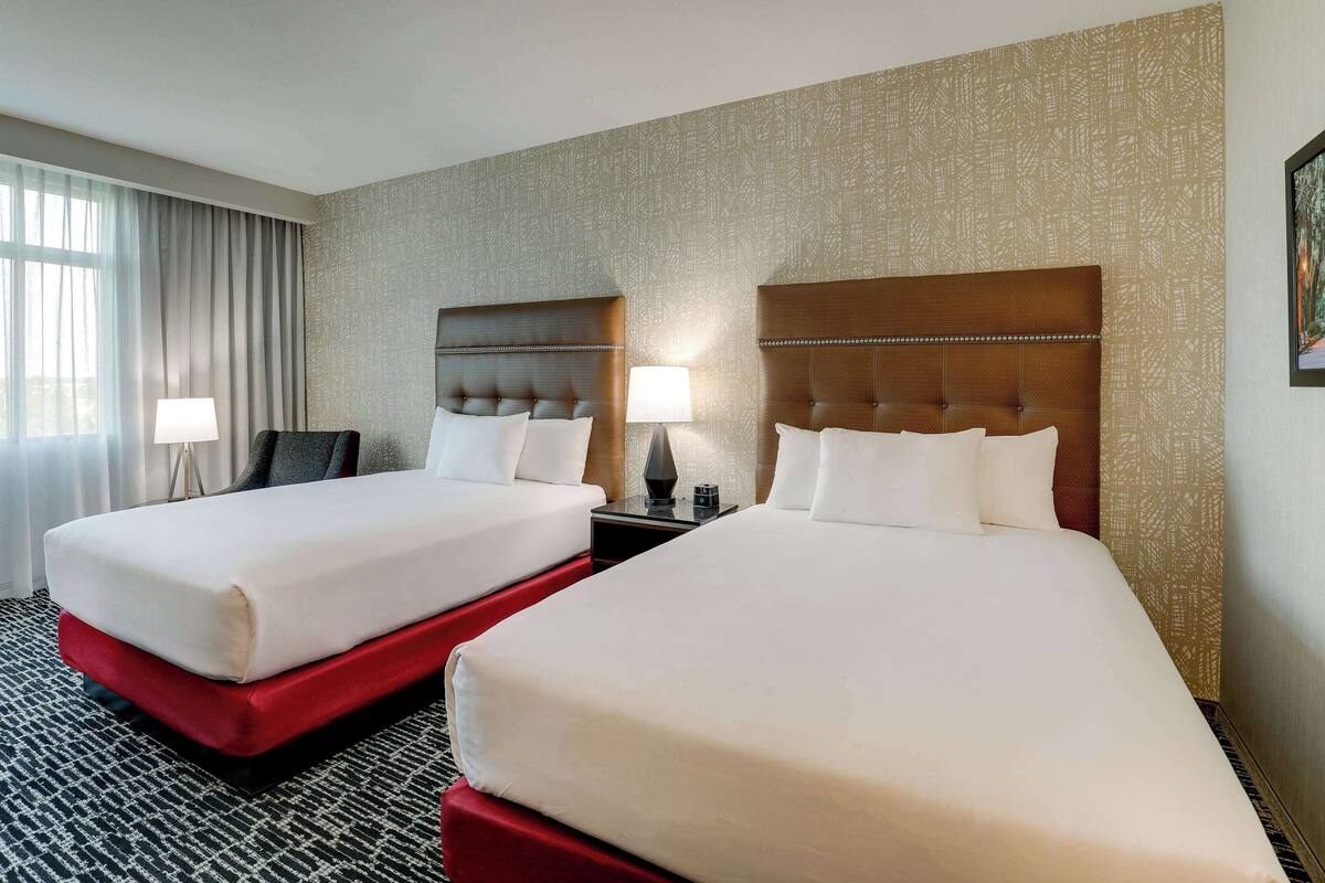 A hotel room with two beds, a red accent bed, a nightstand with a lamp, and a large window with curtains.