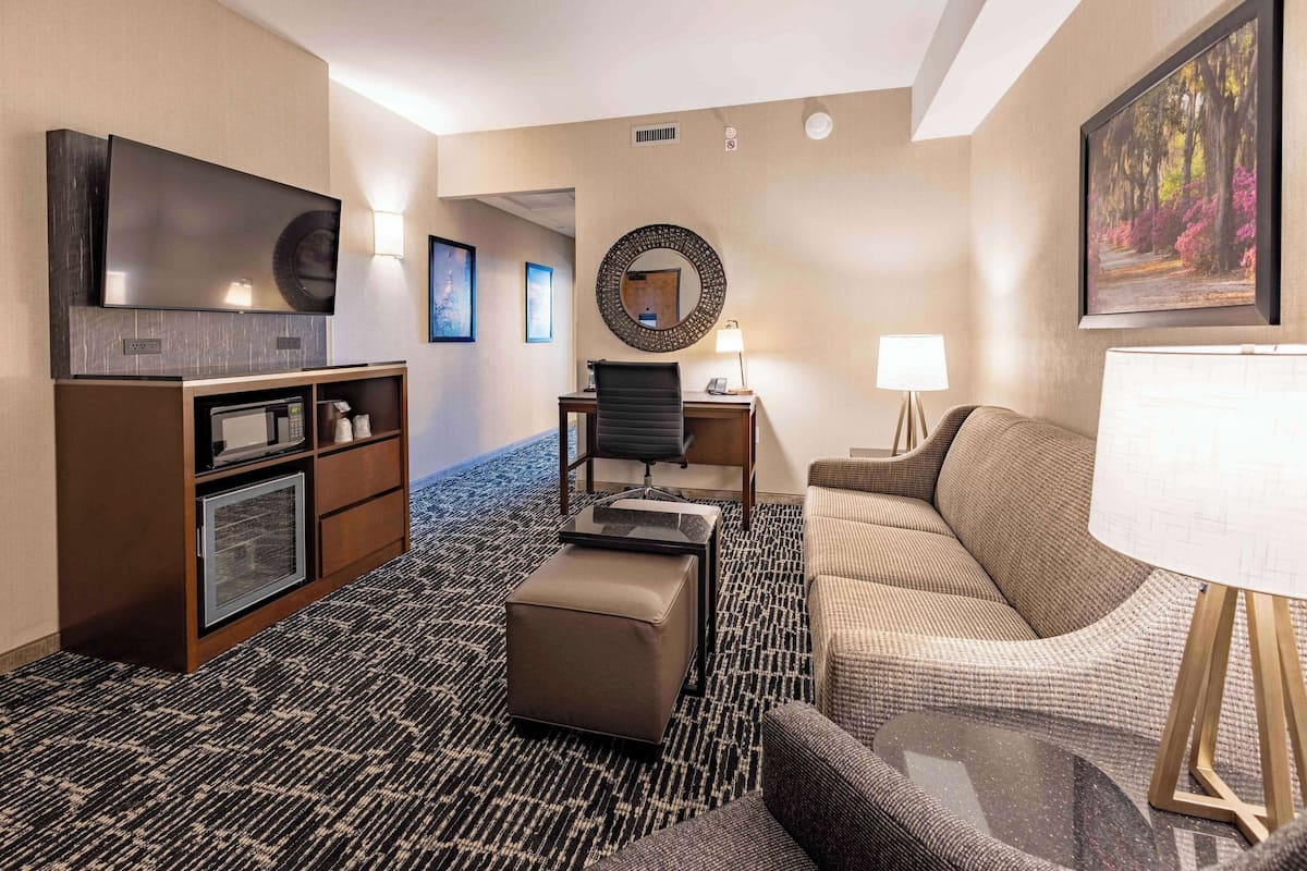 A hotel room with a flat-screen TV, a desk with a chair, a sofa, a round mirror, and a large painting on the wall.