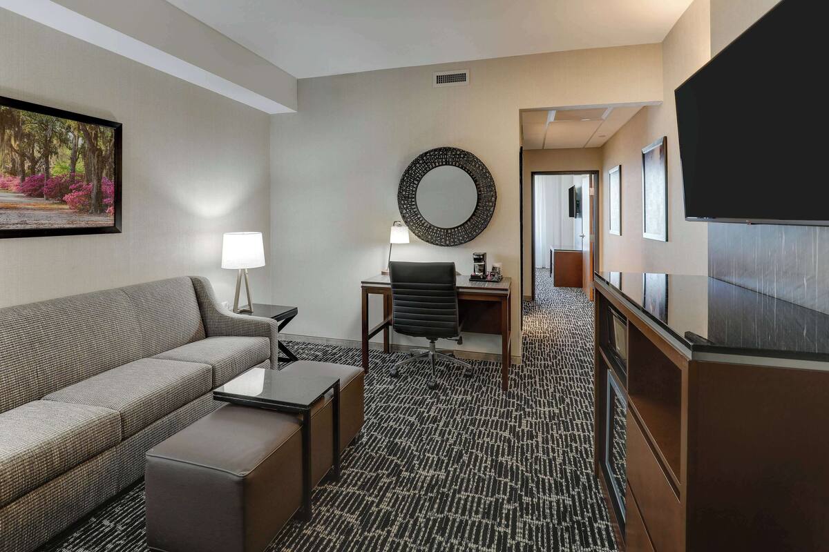 A hotel room with a sofa, a desk with a chair, a TV, a mirror, and a framed picture on the wall.