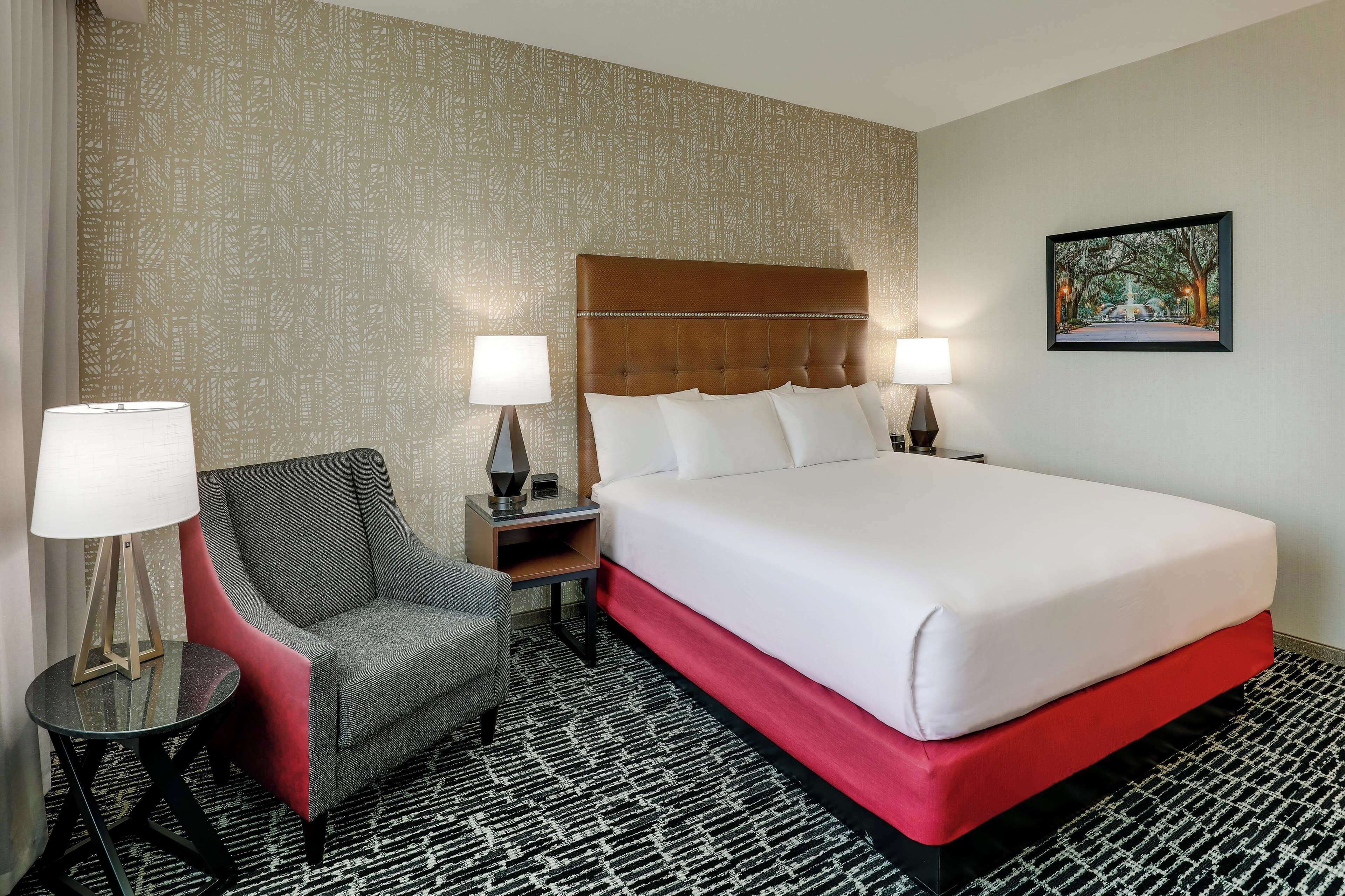 Une chambre d’hôtel avec un grand lit, une chaise, deux tables de chevet avec des lampes et une moquette à motifs.