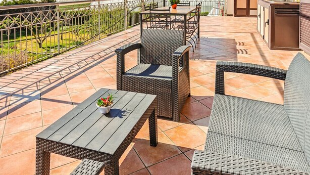 Eine Terrasse im Freien mit Korbmöbeln, einem Tisch mit Topfpflanze und Blick auf einen Garten und Berge im Hintergrund.