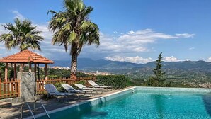 Una piscina con sillones de descanso y un pabellón, rodeada de palmeras y montañas al fondo.