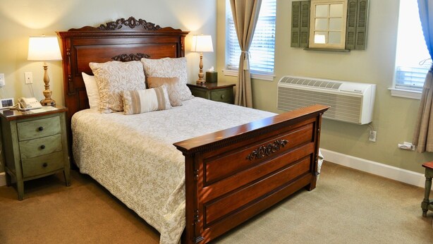 A neatly made bed with a wooden headboard, two bedside tables with lamps, a radiator, and a window with curtains. - Queen downstairs room (cayucos)