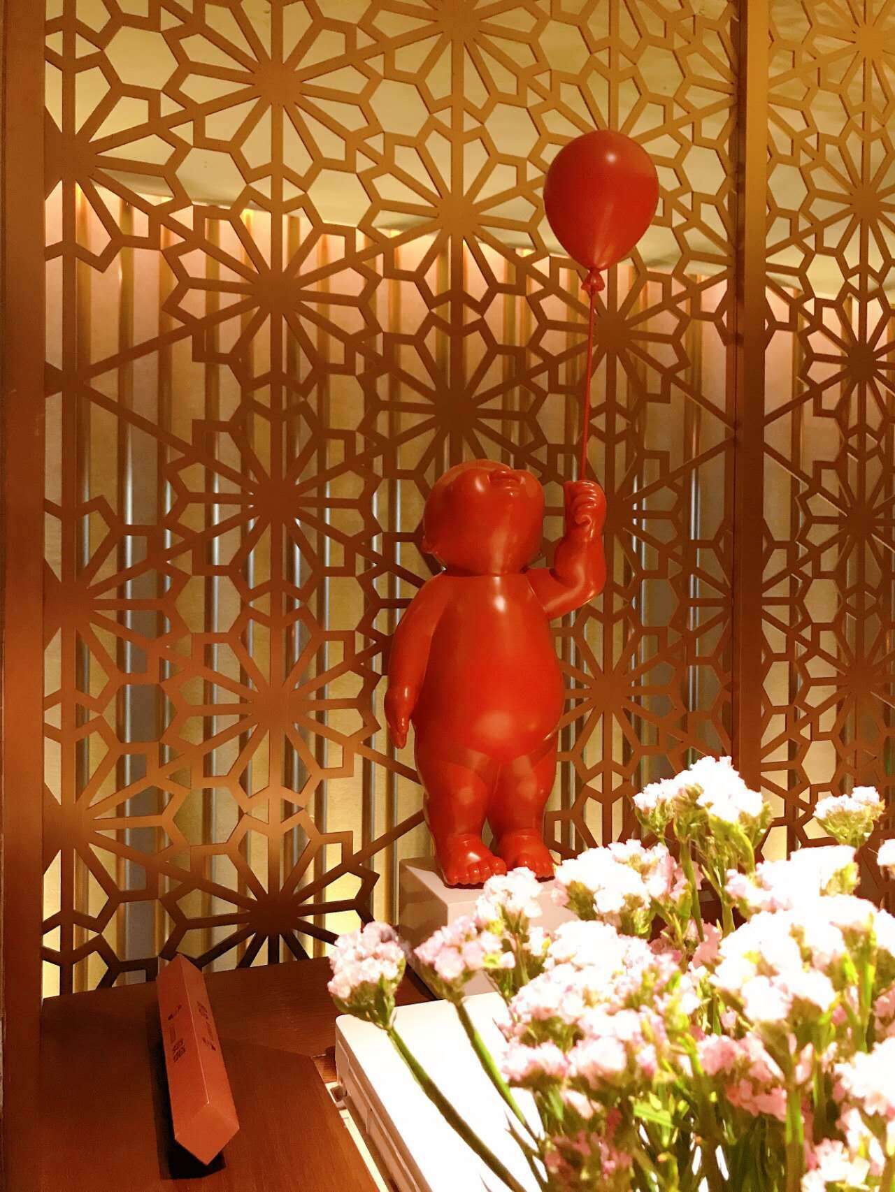 Eine rote Spielfigur eines Kindes, das ein Herz hält, eine weiße Vase mit rosa Blumen und ein Holztisch mit einem roten Gegenstand darauf.