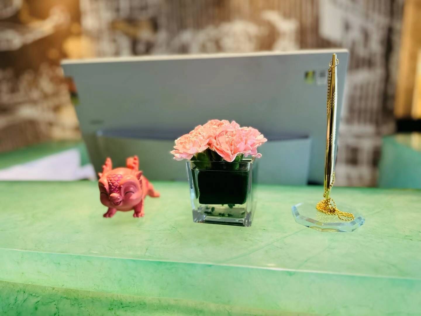 Ett skrivbord med en bärbar dator, en liten vas med rosa blommor, ett guldfärgat pennställ och en liten rosa grisfigur.