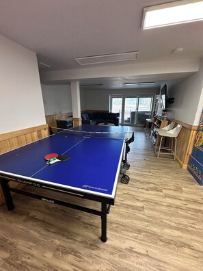 Una mesa de ping pong con superficie azul y red, rodeada por un piso de madera, con una zona de estar y una ventana al fondo.