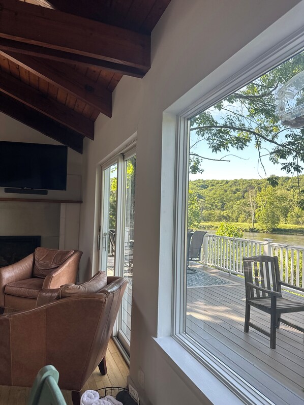A room with a brown leather sofa, a television, and a large window overlooking a deck with outdoor furniture.