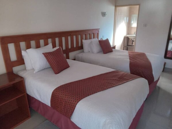 A hotel room with two beds, each with white bedding and red accents, a wooden headboard, and a small bedside table.