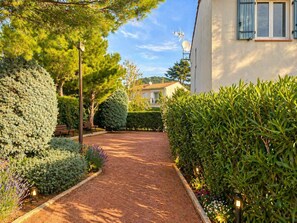 Un chemin menant à un bâtiment surmonté d’une antenne parabolique, entouré de haies et d’arbres bien entretenus.