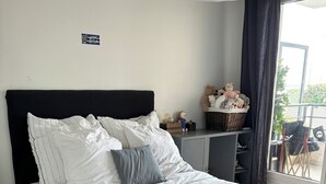 A neatly made bed with white bedding and a dark blue throw blanket. There is a small table beside the bed with a basket and a teddy bear. A large window with a view of the outdoors is visible. - Appartement Lumineux à Suresnes à Côté de Paris Vue Tour Eiffel (Suresnes)