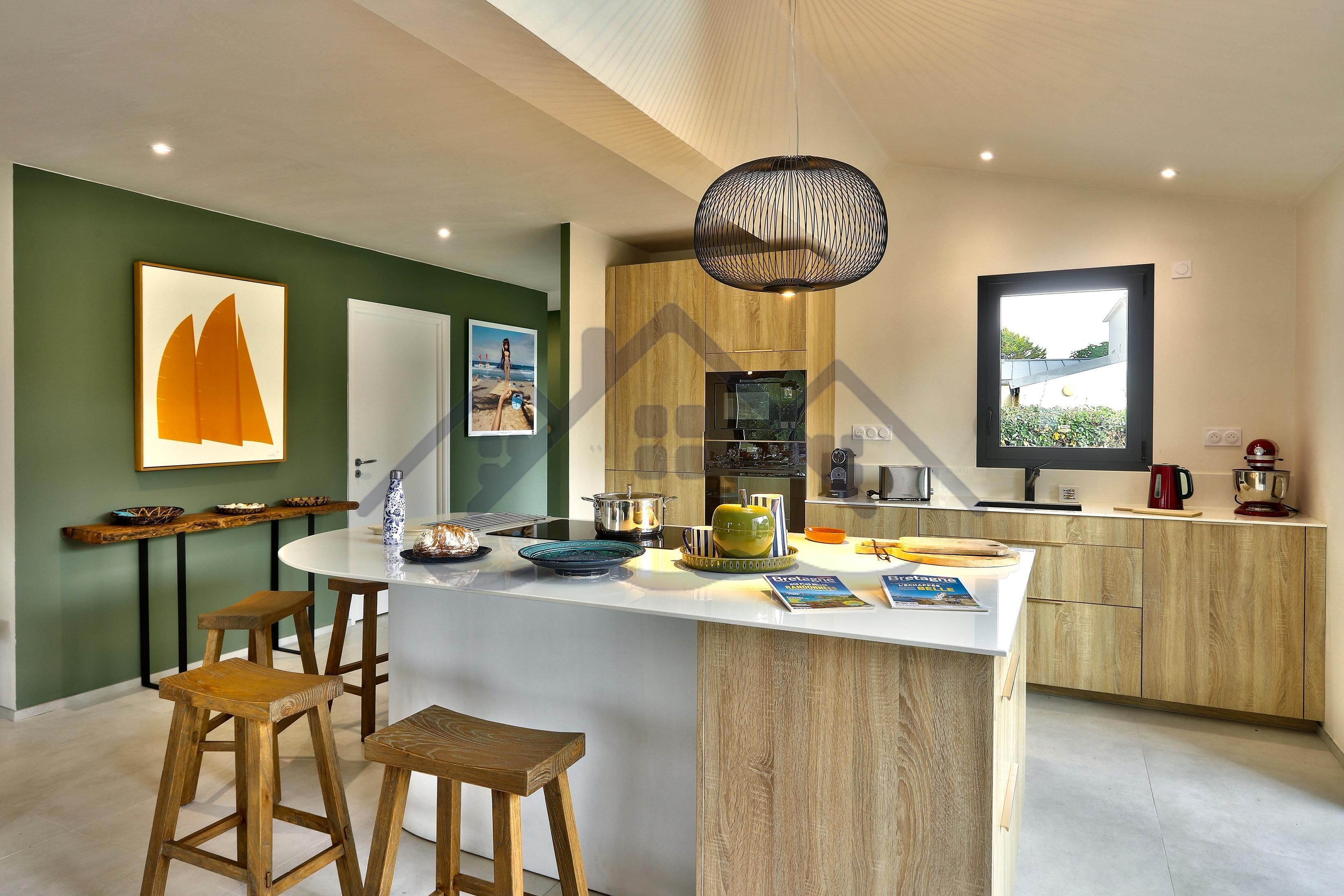 A modern kitchen with a central island, wooden stools, and a hanging light fixture.