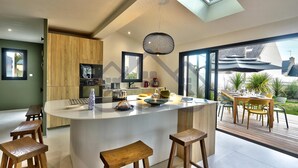 A modern kitchen with a central island, wooden stools, and a dining area with a view of the outdoors. - Large, recently built architect-designed house for 12 people (Guilvinec)