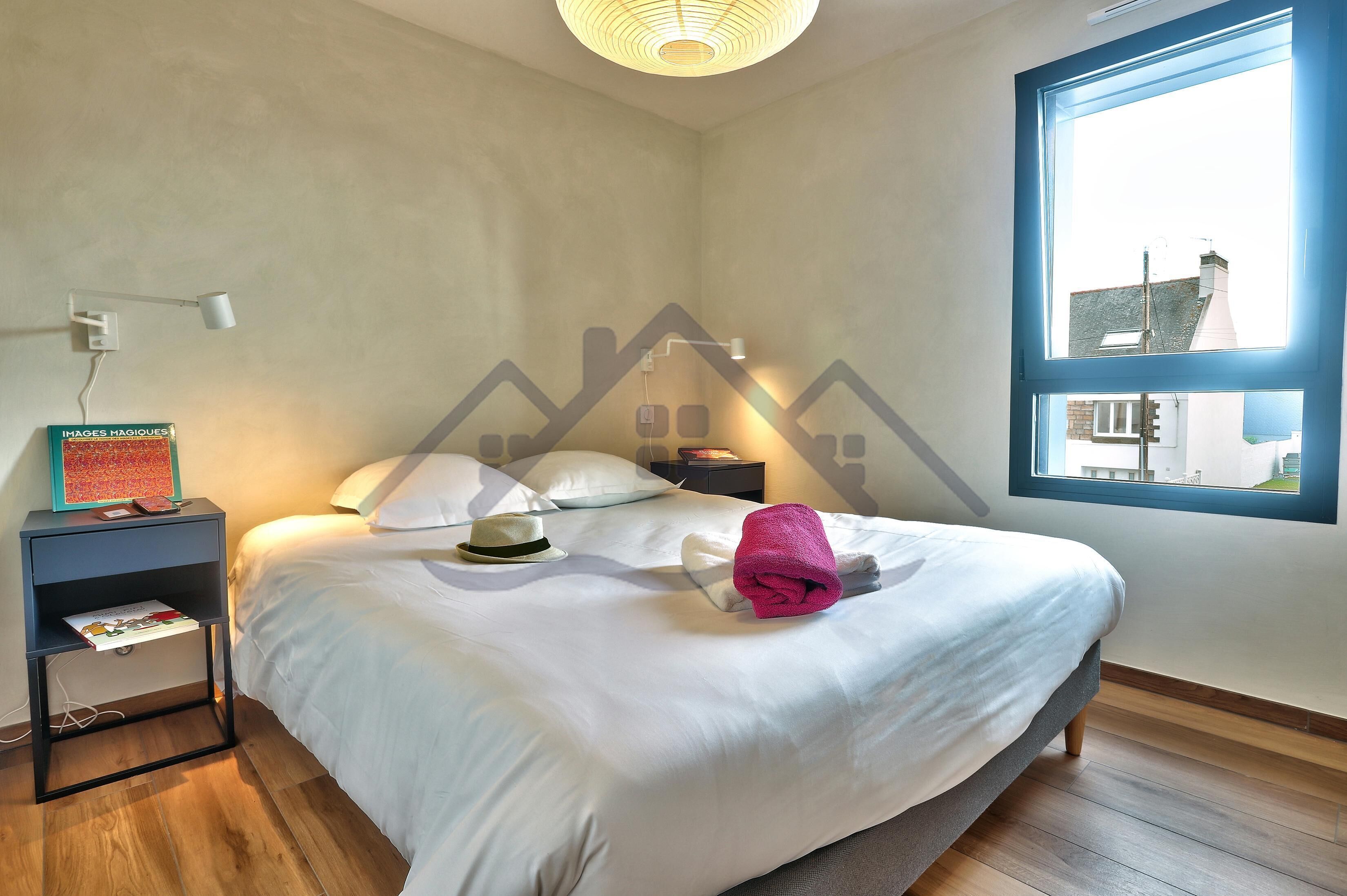 A neatly made bed with a white comforter and a pink towel, a bedside table with a lamp, and a window with a view of buildings.
