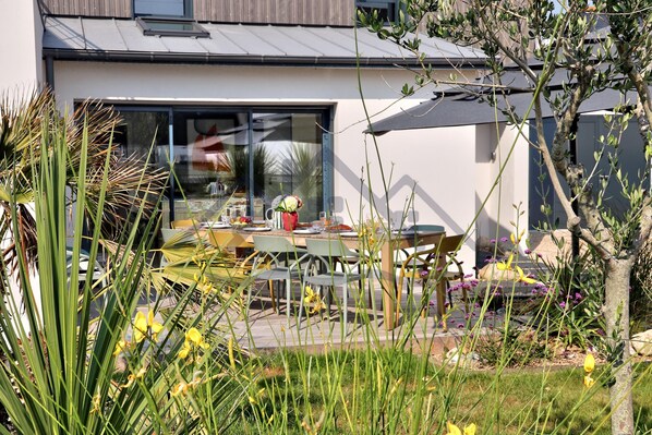 Ett modernt hus med en uteplats som har ett bord och stolar, och som är omgivet av grönska och blommor.