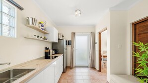 Una cocina con un lavamanos, armarios y una encimera. Hay estantes con objetos y una puerta que da a otra habitación.