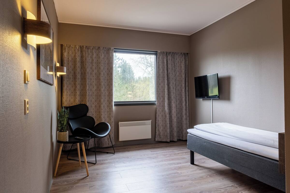 Moderni makuuhuone, jossa on sänky, televisio, tuoli, ikkuna verhoilla ja patteri.