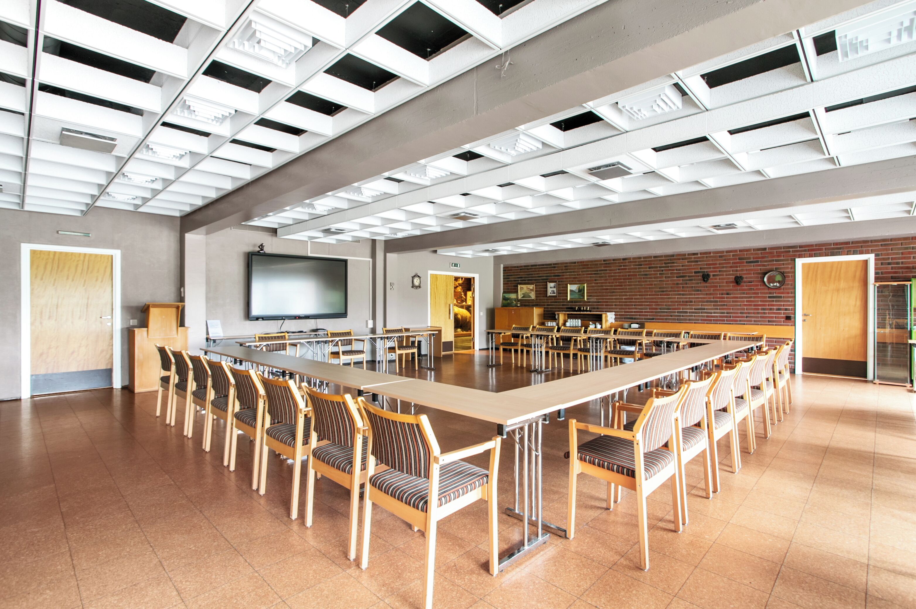 A spacious conference room with a long table and chairs, a projector screen, and a brick wall with framed pictures.
