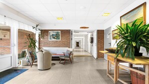 Un hall espacioso con pared de ladrillo, mostrador de recepción, zona de estar con sofá y sillas, y una gran planta en maceta.