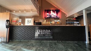 Zona de recepción de hotel con un mostrador de ladrillo negro, un refrigerador de Coca-Cola y un televisor que muestra un fuego.