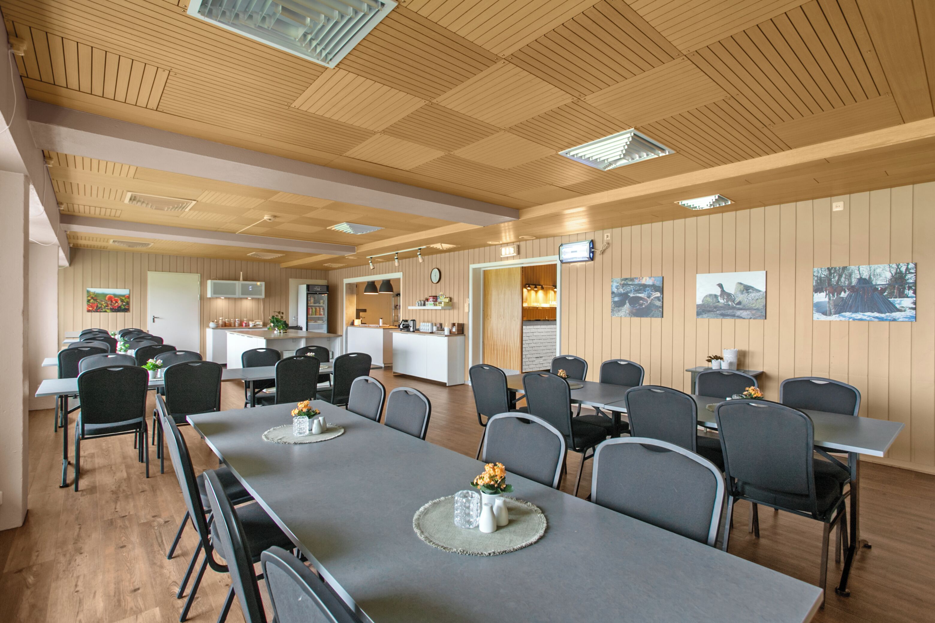 A conference room with long tables and chairs, a kitchen area, and framed pictures on the walls.