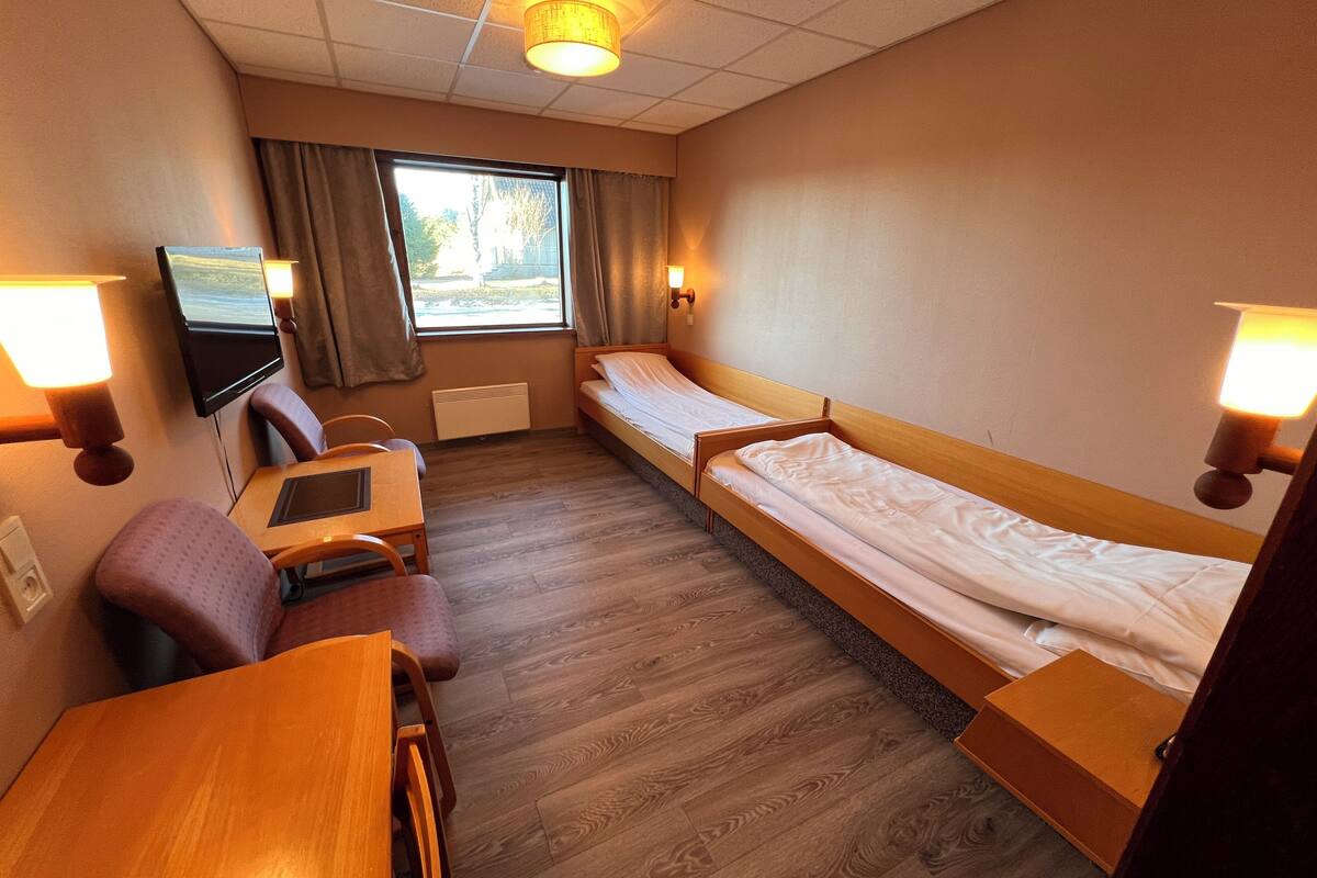 Hotellihuone, jossa on kaksi erillistä sänkyä, työpöytä tuolilla, televisio ja ikkuna, jossa on verhot.
