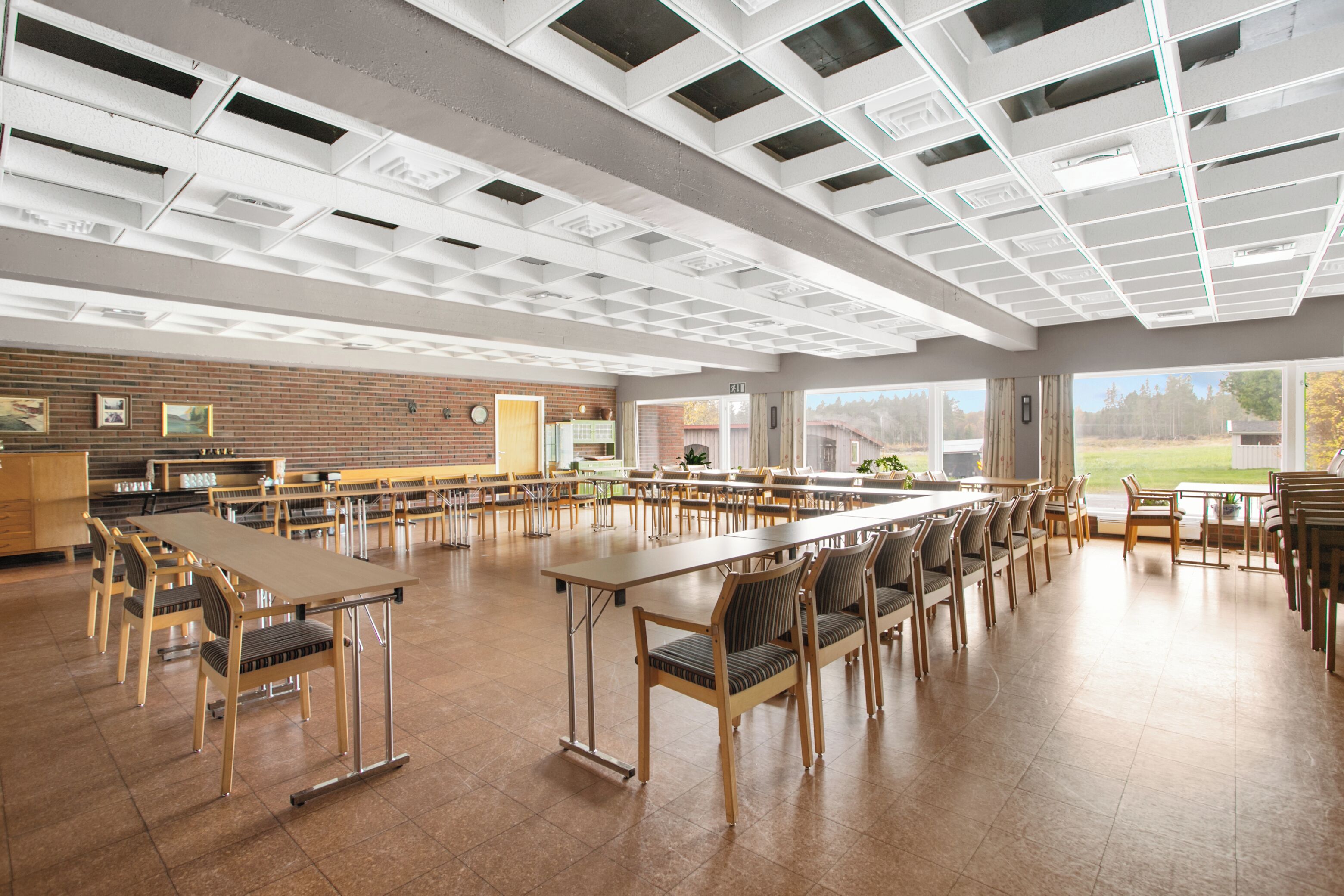 A spacious dining hall with multiple tables and chairs, a brick wall with framed pictures, and large windows offering a view of the outdoors.