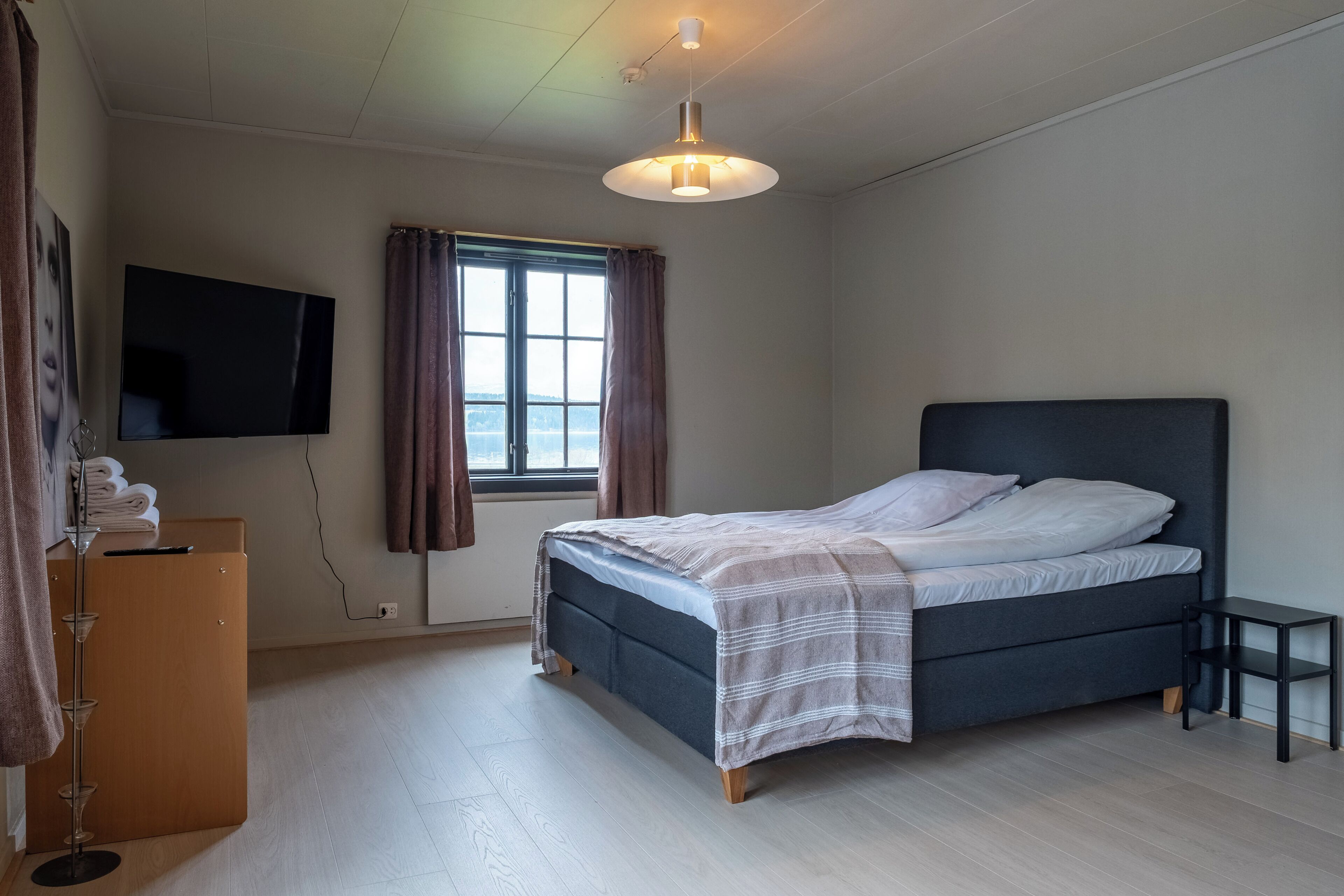 Quarto com cama, televisão na parede, janela com cortinas e mesa de cabeceira.
