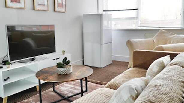 A modern living room with a flat-screen TV, a white refrigerator, a beige sofa, and a small table with a plant. - Clean, Spacious 2-bed Apartment in Brierley Hill (Brierley Hill)