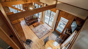 A spacious loft-style room with a high ceiling, wooden beams, a large window, a cozy seating area with a sofa and chairs, and a dining table with chairs.