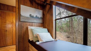 A wooden cabin interior with a bed, a window with a forest view, and a painting on the wall.