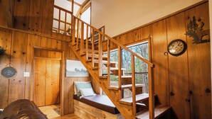 A wooden interior with a staircase, a bed with a pillow, a wooden bench, and a wall clock.