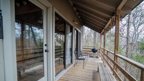 A wooden deck with glass doors leading to a deck area, outdoor seating, and a view of the surrounding forest.