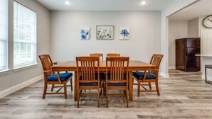A dining room with a wooden table and chairs, three framed floral artworks on the wall, and a clock on the wall.