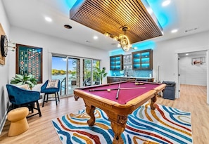 A pool table in a modern room with a large window, a blue chair, and a colorful rug.