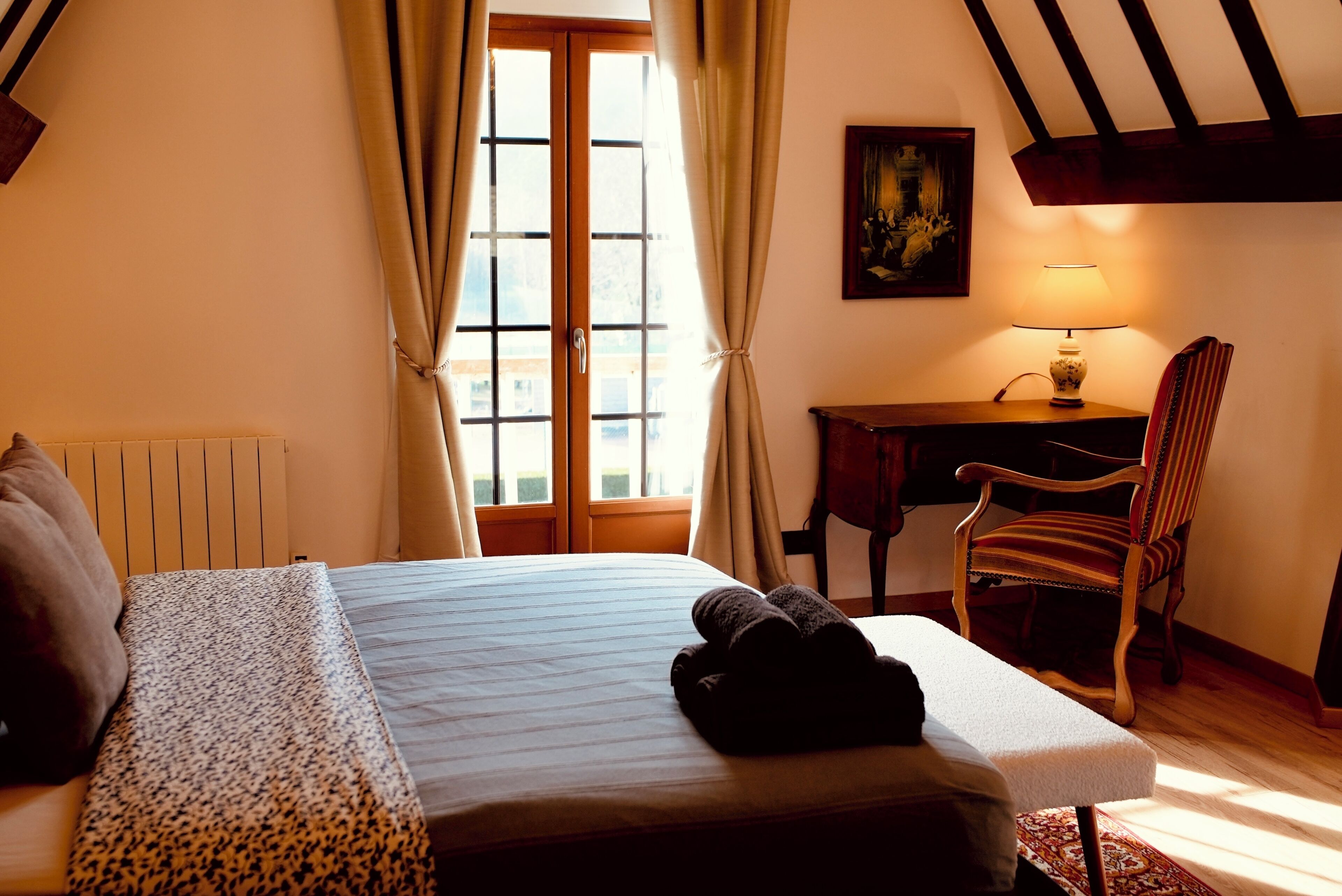 Une chambre d’hôtel avec un lit, un bureau, une chaise, une lampe et une fenêtre avec des rideaux.