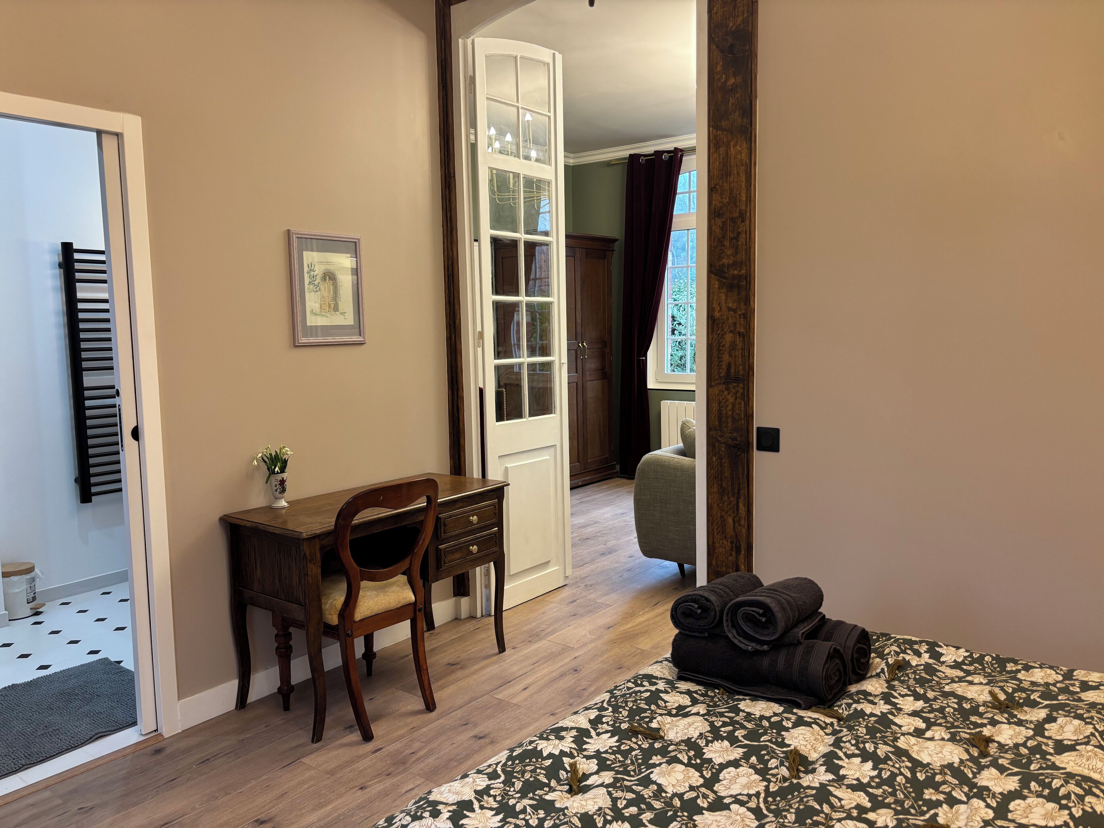 A room with a bed, a desk, and a chair. There is a window with curtains, a radiator, and a door leading to another room.