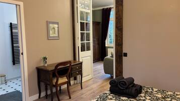 A room with a bed, a desk, and a chair. There is a window with curtains, a radiator, and a door leading to another room.