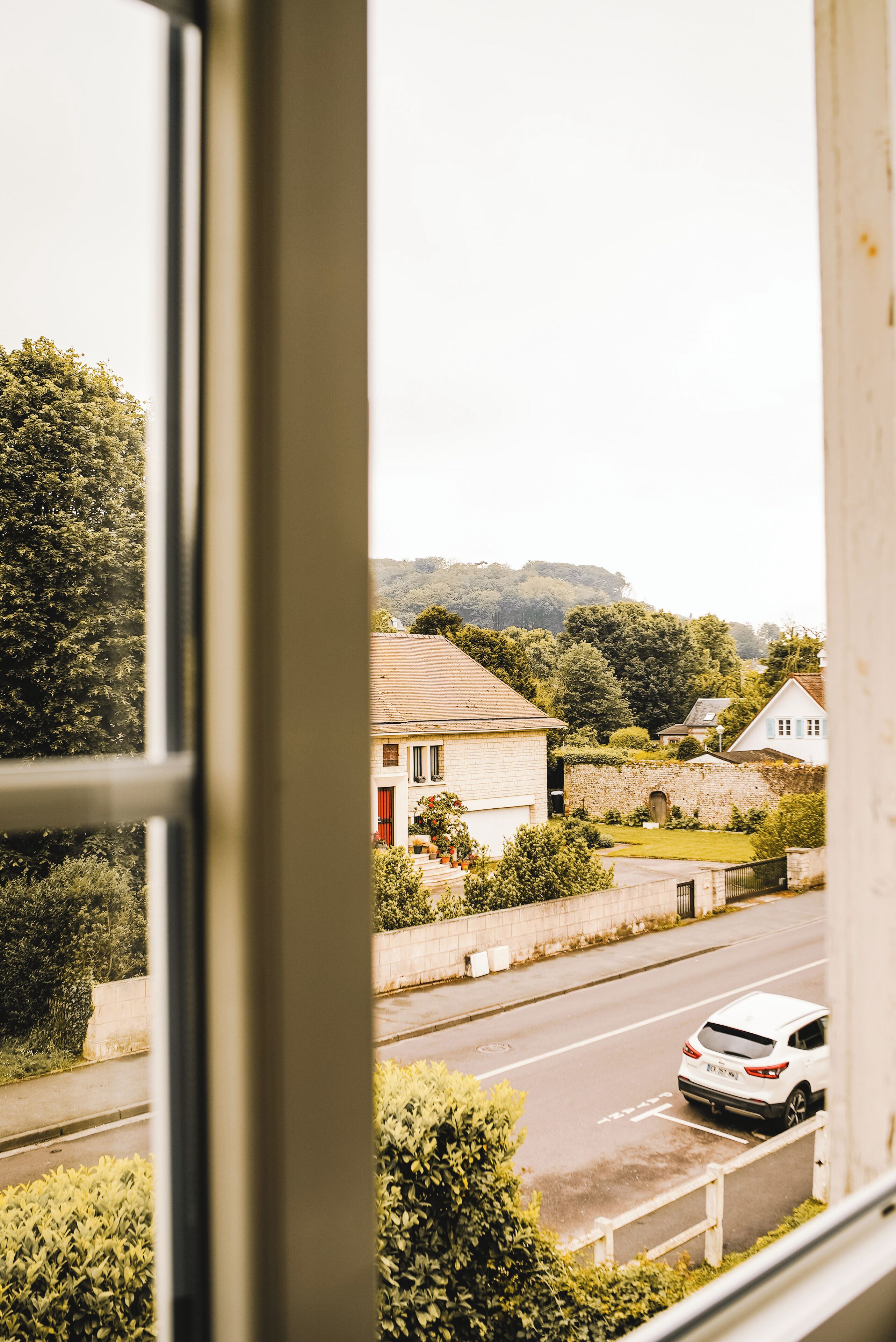 A view through a window of a residential street with houses, a road, and a white car.
