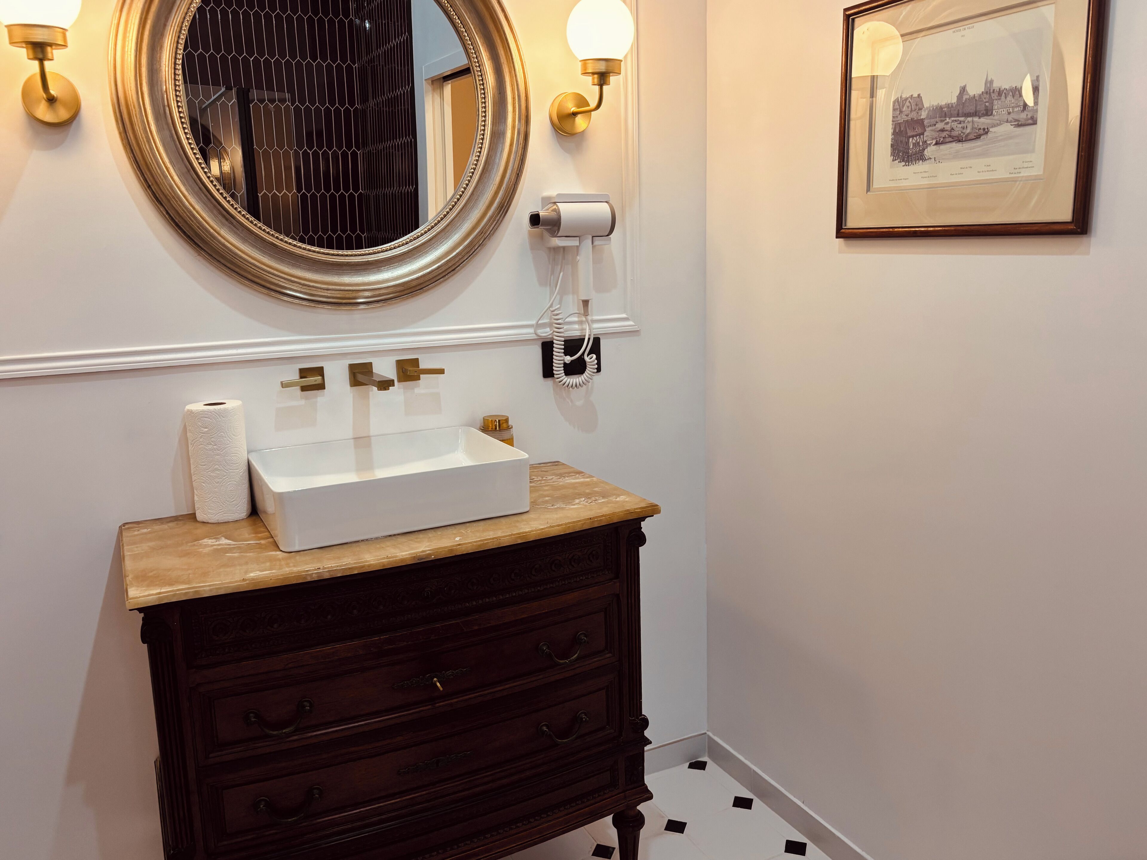 A bathroom with a white sink, dark wooden vanity, gold-framed mirror, and a framed cityscape picture on the wall.
