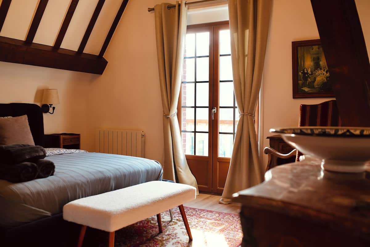A bedroom with a bed, bench, lamp, radiator, window with curtains, and a painting on the wall.