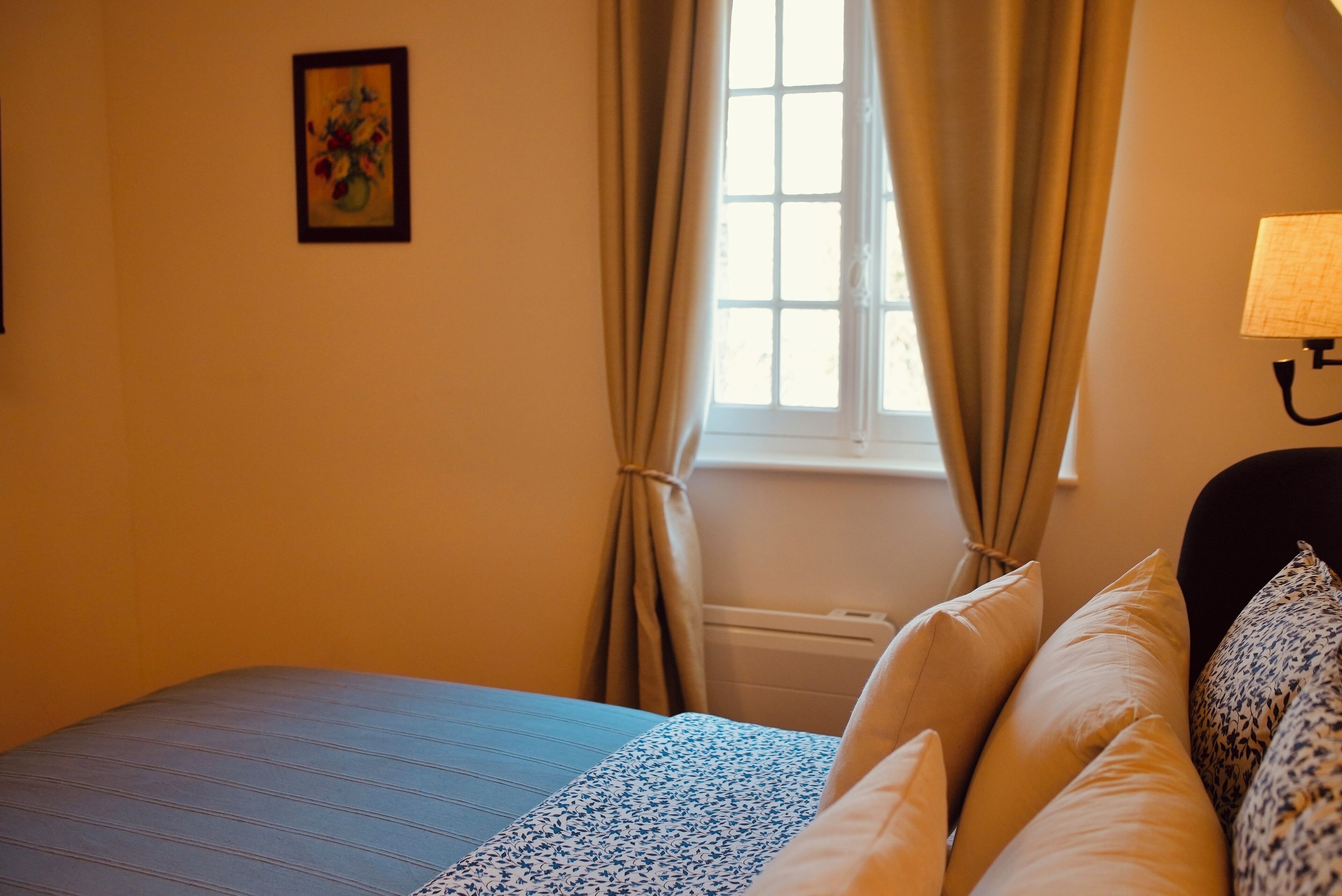 A bedroom with a bed, a window with curtains, a radiator, and a wall-mounted lamp.