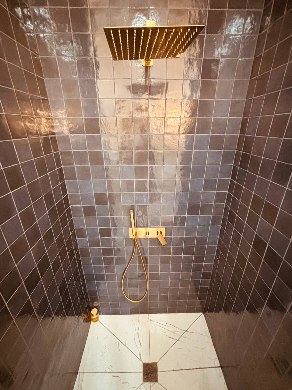 A shower area with a gold showerhead and control panel, tiled walls, and a drain at the bottom.