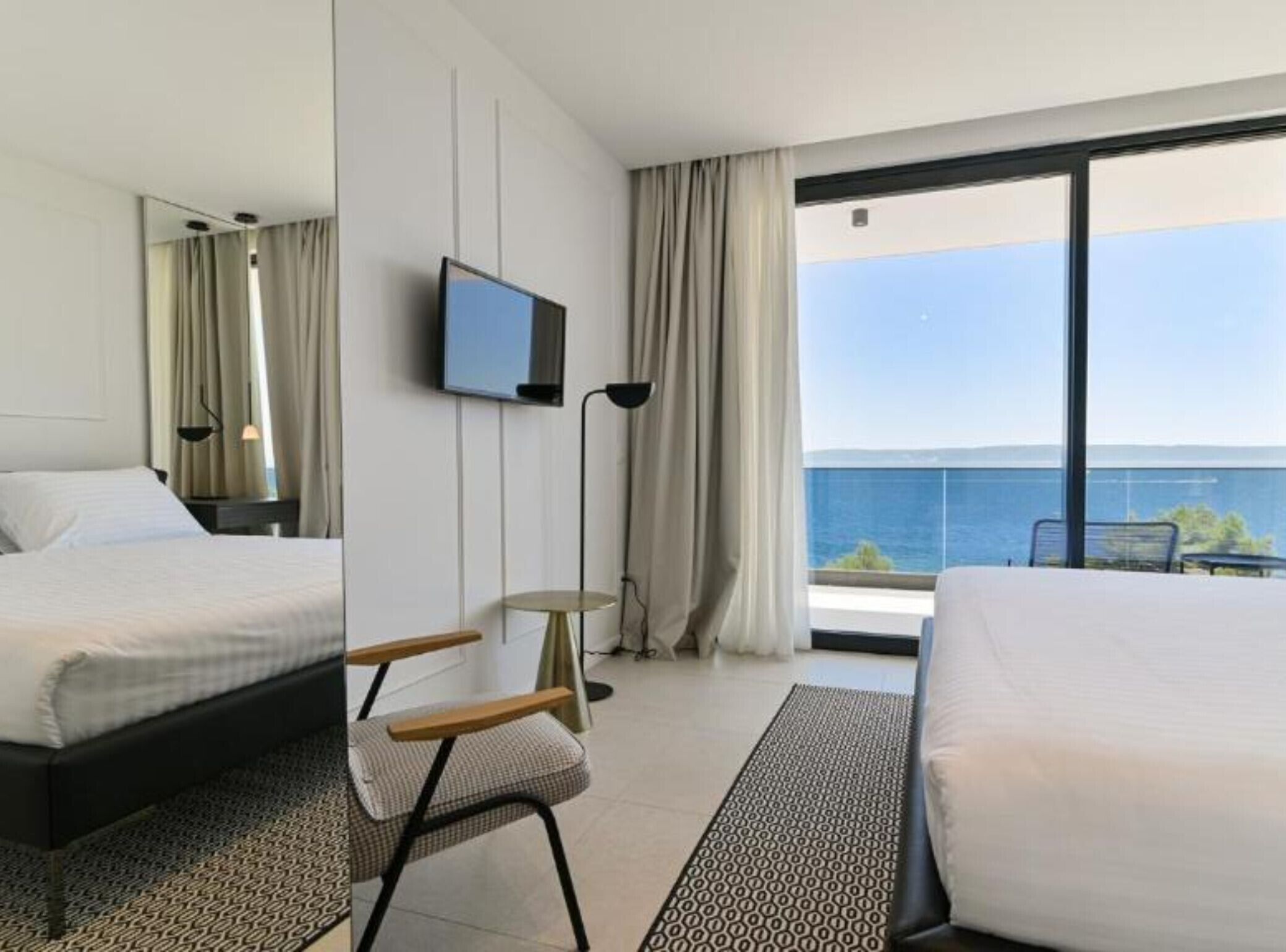 A hotel room with a bed, a TV, a desk with a chair, and a view of the sea.