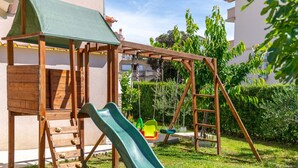 A wooden play structure with a slide and swings in a backyard. - Two Bedroom Apartment (Lopar)