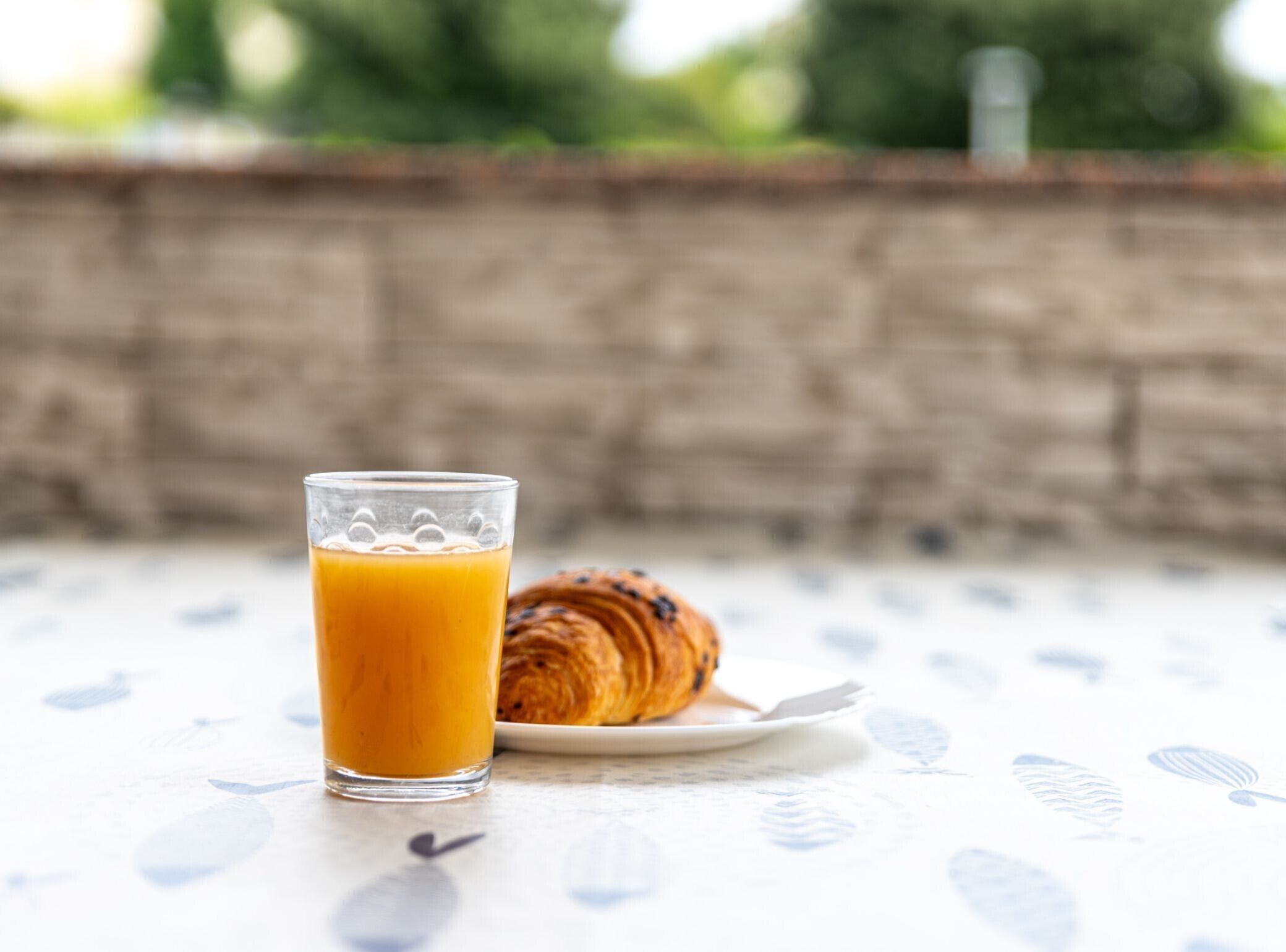 A glass of orange juice and a croissant on a white table with a leaf pattern.