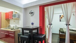 A compact room with a kitchenette, a dining area with stools, a bed with curtains, and a wall clock.
