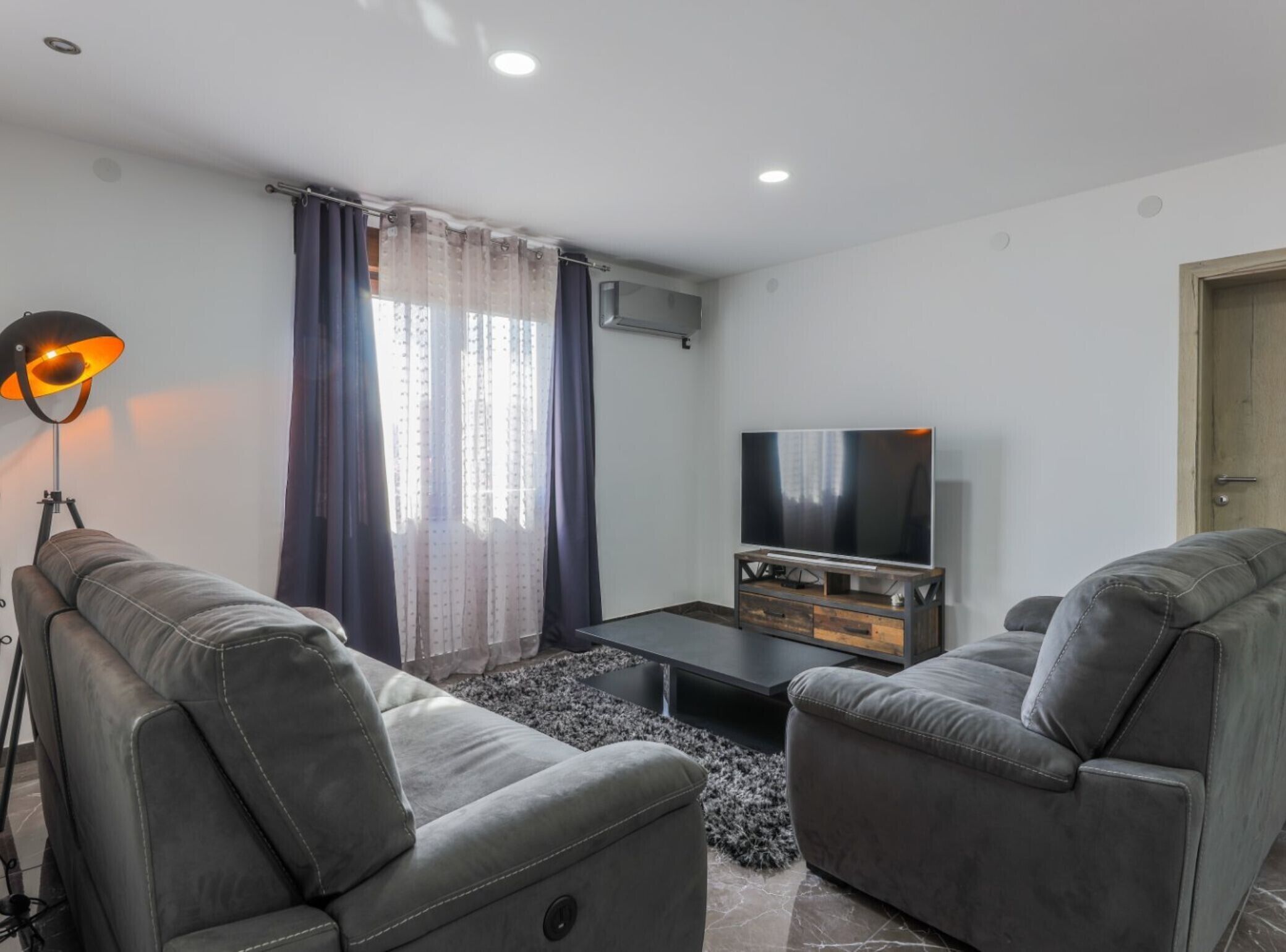 A modern living room with two grey sofas, a coffee table, a TV on a wooden stand, a floor lamp, and a window with sheer curtains.