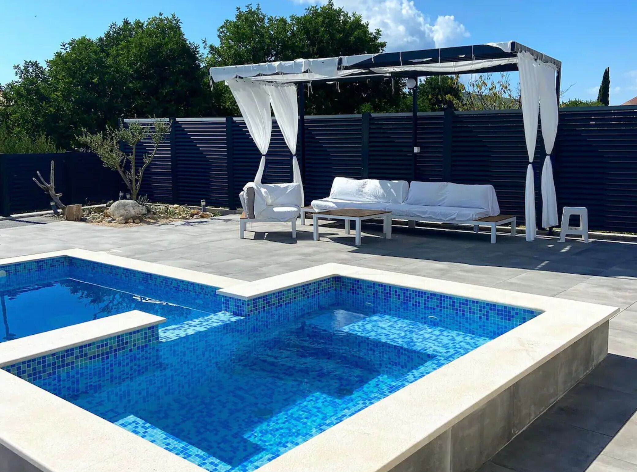 A rectangular swimming pool with blue tiles, surrounded by a tiled deck. There are white lounge chairs and a white canopy structure.